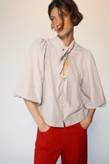 Blusa de cuello subido con escote en pico y cierre frontal con botones forrados. Presenta mangas abullonadas de largo tres cuartos con elástico en los puños y detalle de lazos en el cuello y el frente.