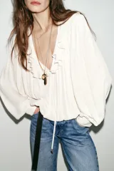 Blusa de georgette en color blanco, con escote en pico, detalle de volantes y lazada frontal. Presenta mangas largas abullonadas con puños ajustados y un bajo asimétrico con diseño tipo globo.