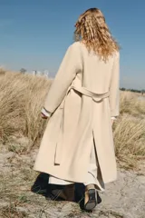 Abrigo largo de lana en color beige, con cuello de solapa, cierre cruzado con botones y cinturón ajustable del mismo tejido. Presenta mangas largas con detalle en los puños y bolsillos de vivo en el delantero.