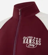Saco femenino de poliviscosa, color blanco con mangas raglán marrones y ribetes blancos. Tiene cuello alto, cierre frontal de cremallera y bordado lettering "Kansas City" en el pecho.