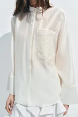 Camisa de cuello mao color beige, confeccionada en tejido satinado con brillo sutil, manga larga con puños anchos y un bolsillo de parche en el pecho con detalles bordados tonales. Cierre frontal con botones ocultos bajo solapa.