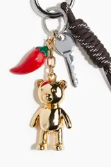 Dije para cartera con llavero de cuerda trenzada marrón y blanco. Incluye dijes colgantes: una llave plateada, un chile rojo esmaltado y un oso de peluche metálico dorado.