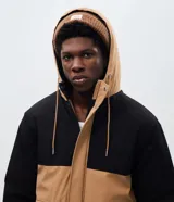 Chaqueta tipo parka de poliéster con diseño en bloques de color negro y tostado. Presenta capucha ajustable con cordones, cierre frontal con solapa superpuesta y bolsillos frontales con tapa.