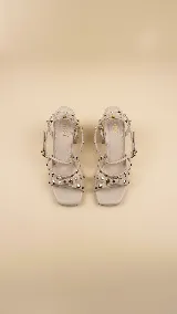 Sandalias de taco ancho de 7 cm, color crema, con tiras cruzadas y cierre de hebilla al tobillo. El diseño presenta múltiples tachas decorativas metálicas en tonos dorados y plateados distribuidas a lo largo de las tiras y en todo el taco.