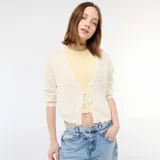Chaqueta de punto con textura de croché en color beige, de corte corto y mangas largas. Presenta un cierre frontal mediante una lazada fina y bordes con acabado festoneado.