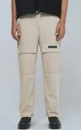 Pantalón cargo de color beige claro, corte recto y holgado, con bolsillos tipo parche en los muslos. Presenta costuras reforzadas en las rodillas y una etiqueta rectangular negra en el bolsillo derecho.