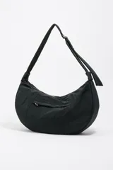Bolso tipo media luna de nylon negro, con correa ajustable para llevar al hombro o cruzado. Presenta costuras verticales que le dan estructura y un pequeño logo de Bimba y Lola en la parte inferior central.