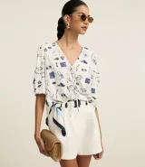 Blusa de mangas abullonadas cortas, confeccionada en Viscolino con 15% de Lino, con cuello v con lazo y estampa de accesorios de playa.