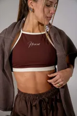 Top deportivo de cuello halter en color marrón chocolate con ribetes en color crema. Presenta el logo de la marca estampado en el centro del pecho.