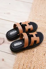 Sandalias planas de cuero sintético, con base negra y tiras anchas en forma de H en color caramelo o negro.