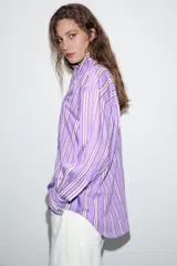 Camisa de corte oversize con diseño de rayas verticales en tonos violeta, blanco y bordó. Presenta cuello subido cruzado con cierre de botones frontal, mangas largas con puños y bajo asimétrico.
