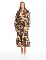 Vestido maxi de crepe con estampado floral en tonos beige y marrón oscuro. Presenta escote en V con botones frontales, mangas largas amplias con puños abotonados y cintura ajustable mediante cordón trenzado. La falda cuenta con un diseño de vuelos escalonados que aporta movimiento.
