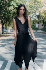 Vestido midi de viscosa color negro, sin mangas, con escote en pico adornado con encaje a tono. Presenta un detalle de nudo drapeado en la cintura y bajo asimétrico.