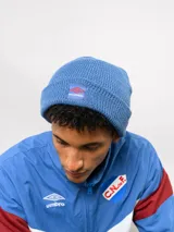 Gorro tipo beanie de tejido de punto color blanco crema, con parche frontal que incluye el logo de Umbro y la inscripción Nacional.