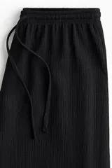 Pantalón negro de tiro alto, confeccionado en tejido texturizado con corte holgado y piernas amplias. Cuenta con cintura elástica fruncida y cordón ajustable.
