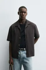 Camisa de manga corta con cuello bowling y calce relaxed fit, confeccionada en tejido seersucker elástico con textura. Presenta un diseño a rayas verticales en tonos oscuros y cierre frontal con botones.
