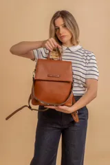 Mochila tipo cartera con tapa, confeccionada en símil cuero color suela. Cuenta con doble compartimento, división interna, cierre de seguridad y bolsillo trasero con cierre. Incluye una correa frontal decorativa con tachas metálicas.