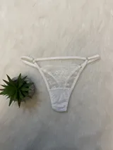 Conjunto de lencería de encaje blanco compuesto por un bralette triangular, una tanga con tiras y un portaligas integrado. El diseño presenta detalles de encaje floral y breteles regulables.
