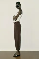 Pantalón tobillero de tiro medio, color beige, con pinzas delanteras y bolsillos frontales. Incluye un cinturón fino de cuero marrón con hebilla metálica dorada y cierre frontal con cremallera, botón interior y gancho.
