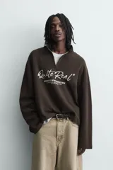 Sudadera de corte boxy fit en tejido de algodón con textura acanalada, color marrón oscuro. Presenta cuello subido con cierre frontal de cremallera y estampado frontal con texto en contraste.