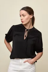 Blusa de manga corta color negro, con calce relajado, escote redondo con abertura y lazo para anudar. Presenta detalles de tablas horizontales en la parte superior que aportan textura al diseño.