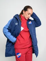 Parka larga de abrigo Umbro del Club Nacional de Football, color rojo con detalles en naranja en las mangas. Cuenta con capucha, cierre frontal con botones a presión y escudo del club bordado en el pecho.