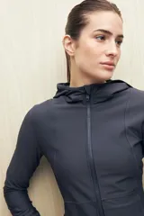 Campera deportiva negra de calce entallado, con cierre frontal completo, capucha integrada y mangas largas con puños envolventes. Presenta costuras estratégicas que estilizan la silueta y bolsillos laterales funcionales.