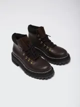 Botas acordonadas de estilo montañés, confeccionadas en cuero marrón oscuro con detalles en gamuza al tono. Presentan una suela track robusta de goma negra con costura visible y ganchos metálicos para los cordones.