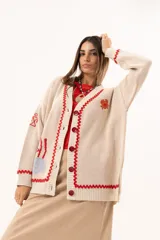 Saco tipo cardigan oversize de punto color beige, con diseño temático marino que incluye bordados de corales, peces, estrellas de mar y tortugas. Presenta detalles de galón picot en color rojo en los bordes y puños, cierre frontal con botones al tono y corte holgado.