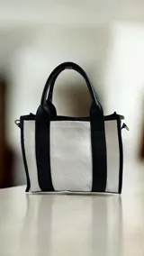 Cartera de cuero modelo Tali, de diseño estructurado que combina los colores blanco, negro y dorado, con detalles de arte pintado a mano en el frente. Cuenta con doble asa de mano, correa larga desmontable y herrajes metálicos.