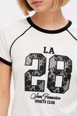 Remera de algodón blanca de manga corta estilo ringer, con cuello y puños en contraste negro. Presenta un estampado frontal con el número '28' relleno de encaje y la inscripción 'San Francisco Sports Club'.