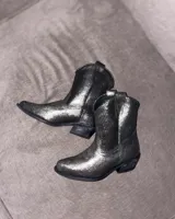 Botas estilo tejano de cuero vacuno con acabado brillante en tono grafito. Presentan un diseño clásico con costuras decorativas en el empeine, caña media y taco bajo.