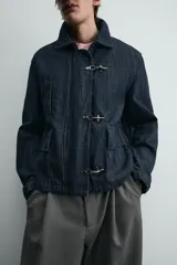 Chaqueta de denim de algodón en color azul oscuro, con cuello solapa y manga larga. Presenta cierre frontal con cremallera oculta bajo una solapa con alamares metálicos, bolsillo de vivo en el pecho y bolsillos de plastrón con solapa en la cadera.