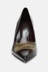 Zapato de tacón alto fino con acabado en punta. Presenta un aplique metálico dorado en la parte frontal y un acabado de textura brillante en color marrón oscuro.