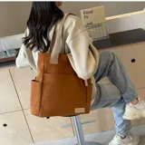 Cartera tipo shopper de lona color marrón, con amplio compartimento principal, bolsillos exteriores laterales y frontales, y asas de hombro en cinta resistente.
