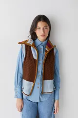 Chaleco de lana con cierre frontal completo y cuello alto. Presenta un diseño de bloques de color en tonos marrón chocolate y celeste, combinando secciones de tejido trenzado con secciones de tejido acanalado. Posee un corte asimétrico, siendo más largo en la parte posterior, y aberturas laterales en el ruedo.