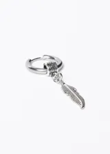 Aro de acero quirúrgico plateado con dije colgante de pluma tallada. El aro es de tipo argolla con cierre a presión y tiene un pequeño adorno con diseño griego. El dije de pluma es alargado y delgado, con detalles grabados que simulan las barbas de la pluma.