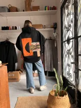 Campera de paño color negro con capucha y diseño de collage en la espalda. El collage incorpora parches en tonos naranja, verde oliva y un tejido con patrón geométrico étnico.