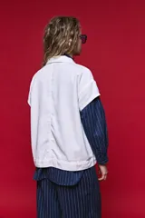 Chaleco de jean color blanco, de corte oversized, con cuello camisero, bolsillos delanteros con solapa y cierre frontal por botones metálicos.