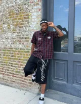 Conjunto compuesto por una camisa de manga corta a cuadros rojos y negros con detalles gráficos bordados y un short negro de corte holgado con costuras en contraste y estampa lateral con tipografía cursiva.