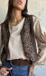 Chaleco de gamuza color chocolate con bordados decorativos en hilo claro y detalles de tachas metálicas en el frente. Presenta cierre frontal con botones forrados.