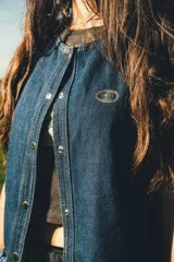 Chaleco de denim azul oscuro con costuras contrastantes en hilo ocre, cierre frontal con broches metálicos a presión y detalle de bordado ovalado en el pecho.