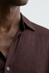 Camisa de manga larga color marrón oscuro, con cuello solapa y cierre frontal de botonadura. El tejido es semitransparente y presenta una textura jacquard.