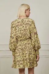 Vestido corto color amarillo con estampado abstracto en marrón, escote en V cruzado, mangas largas abullonadas con puños elásticos y falda con volantes.