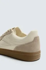 Zapatilla deportiva de estilo retro con diseño de paneles combinados en tonos beige y crema. Presenta detalles en gamuza y una suela de goma en color caramelo.