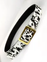 Cinturón de cuero genuino con estampado de manchas tipo vaca en blanco y negro. Presenta una hebilla rectangular de metal en tono bronce envejecido y forro interior negro.