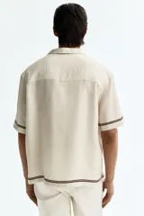 Camisa de manga corta color beige, con cuello abierto y cierre frontal de botones. Presenta un estampado en marrón de palmeras, soles y estrellas, con líneas en los bordes.