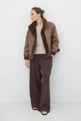 Chaqueta de estilo aviador en color marrón, confeccionada en material tipo gamuza con interior y bordes de piel sintética al tono. Presenta un corte holgado, mangas largas y cierre frontal.