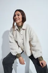 Campera de corderito color beige, con cuello camisero y cierre frontal de botones a presión. Presenta una cartera central lisa en contraste y mangas largas con puños elásticos.