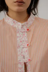 Blusa celeste a rayas blancas, de manga corta abullonada. Cuenta con cuello mao y tapeta de botones con detalles de estampado floreado en tono rosado, presentes también en el cuello y los puños.
